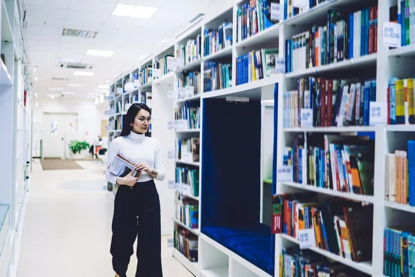 在当代明亮的图书馆里 身穿眼镜和休闲装 头戴五颜六色书籍的年轻而专注的亚洲女性走在现代书架旁边 — 图库照片