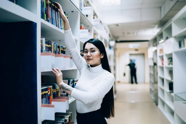 迷人的年轻亚洲女性穿着白领衬衫和眼镜 站在大学图书馆的书架上 从书架上取书 — 图库照片