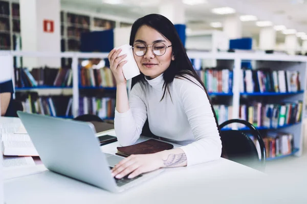 若いです眠いアジアの女性学生で眼鏡をかけコーヒーカップを保持し ノートパソコンで作業しながら 図書館の机に座って — ストック写真