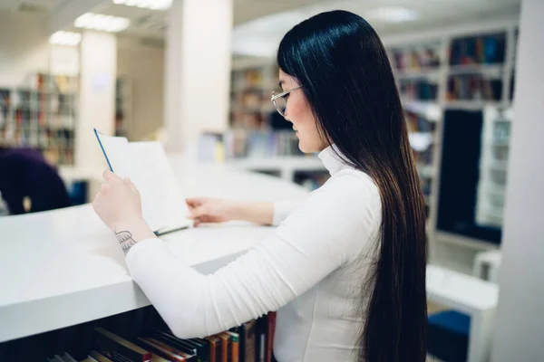 図書館の白いカウンターに立つ興味のある若い女性の側面図と目次を読む — ストック写真