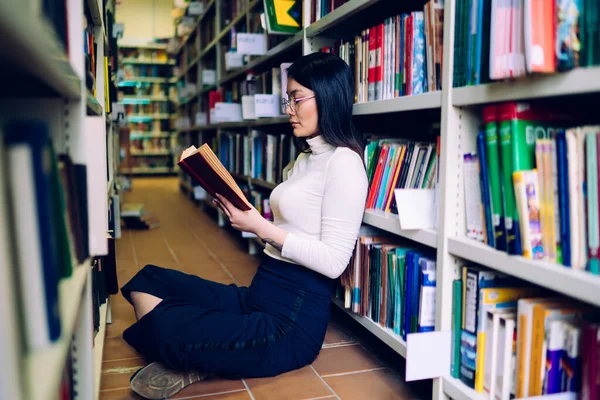 在图书馆的书架和阅读书籍之间 年轻漂亮而专注的女学生双腿交叉坐在地板上的侧视图 — 图库照片