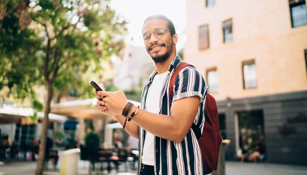 町で晴れた日の間にナビゲーションのためのスマートフォンを使用して陽気な暗い肌の男性旅行者の肖像画 笑顔アフリカ系アメリカ人ヒップスター男は屋外で過ごす時間をチャットでメッセージを送信 — ストック写真