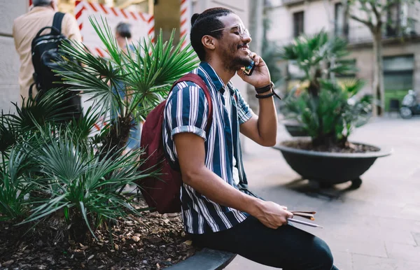 Smiling african american hipster guy traveler resting during journey and talking on mobile phone via international connection, positive dark skinned male tourist using roaming for making call