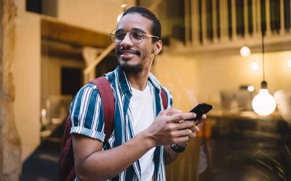 Happy positive african american man holding mobile phone sending text messages and looking away, positive handsome dark skinned hipster guy communicating via smartphone and 4G connection