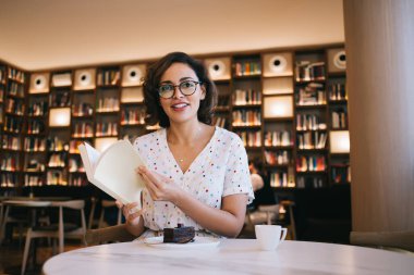 Gözlüklü bir kadın kütüphanede dinlenirken masada kitap okuyup kameraya bakıyor.
