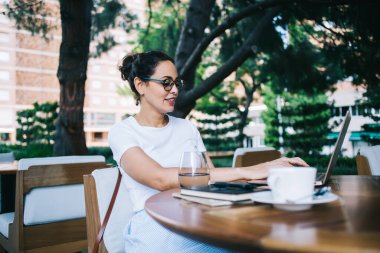 Gündüz vakti uzaktan çalışırken, dizüstü bilgisayarı ve bir bardak suyla masada oturan, günlük kıyafetleri ve gözlükleriyle genç ve zeki bir kadın.