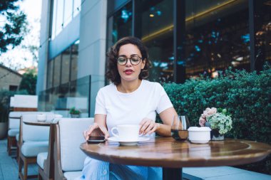 Modayı takip eden kısa boylu, gözlüklü çekici bir kadın modern şehir bölgesinde açık büfe masasında taze demlenmiş kahveyle oturuyor.