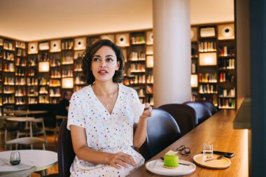 Genç, düşünceli bir kadın, masada lezzetli kremalı pastayla birlikte kitapçının kafeteryasına bakıyor.