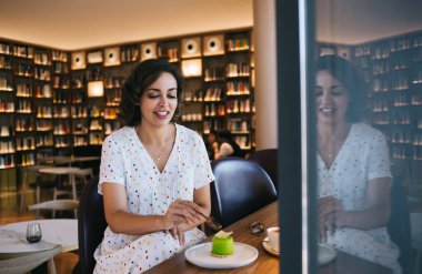 Kaşıkla gülümseyen genç bir kadın kitapçının kahve dükkanındaki masada bir fincan kahvenin yanında lezzetli bir kek yemeye hazırlanıyor.