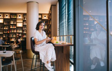 Kafeteryada bacak bacak bacak üstüne atmış genç bir kadın pasta ve içecekle otururken cam duvara yansıyor ve kameraya bakıyor.