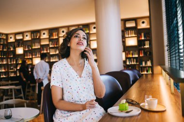 Genç hayalperest kadın cep telefonuyla konuşuyor. Yanında tatlı ve kahve var. İsimsiz insanların yanında.