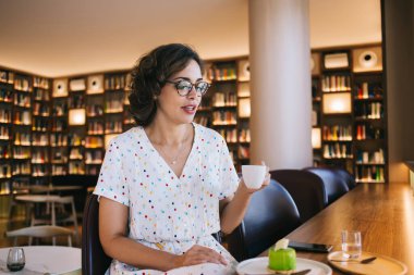 Gözlüklü genç bayan bir bardak içecekle kitapçının kafesindeki lezzetli kremalı pastaya bakıyor.