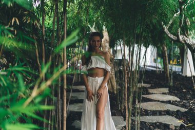 Bambu ormanında dikilip güneşli bir yaz gününün tadını çıkarırken kameraya bakan beyaz bluzlu, cılız bir kadın.