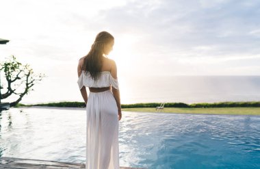 Tanımlanamayan beyaz elbiseli bir kadının gölün kenarında dikilip güneşli bir sahilde yaz tatilinin tadını çıkarışının görüntüsü.