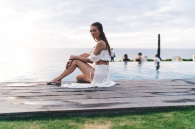 Bütün vücudu çıplak ayakla dalgın beyaz bir kadın ahşap havuz kenarında oturuyor ve dinlenirken kameraya bakıyor.