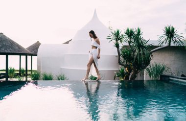 Yüzme havuzunun kenarında duran ve havuz kenarına bakarken bacağıyla suya dokunan tanınmayan bayanın yan görüntüsü.