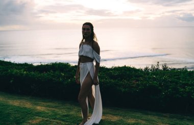 Gün batımında dalgalı denizin yanındaki çimenli tepede kameraya bakarken tanınmayan uzun saçlı bir kadın.