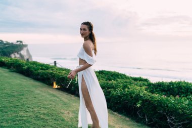 Yeşil sahilde gezinirken omuzlarının üzerinden kameraya bakan modaya uygun beyaz elbiseli muhteşem bir kadın.