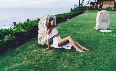 Yüksek açılı, çıplak ayaklı, sakin, zarif elbiseli bir kadın yeşil çimenlere yaslanırken kameraya bakıyor.