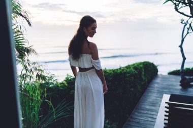 Beyaz etekli, ince, çekici bir kadın. Terasta dikilip arkasını kolluyor.