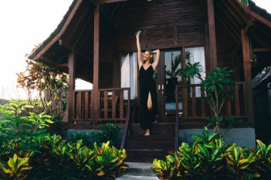 Cep telefonu olan iyimser bir kadın yaz tatilinde tatil köyünde yeşil yaprakların arasında geleneksel kır evinin girişinde duruyor.