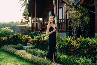 Yeşil yoldaki keyifli bayan yaz tatilinde yemyeşil bitkilerle dolu ahşap evin yanında akıllı telefonuyla geziyor.