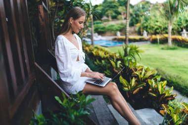 Pozitif odaklanmış beyaz elbiseli genç bir kadının tropikal bahçedeki ahşap merdivenlerde oturup uzaktan bilgisayarla çalışmasının yan görüntüsü.