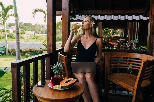 乐观的女性自由职业者一边坐在餐桌旁 一边端着新鲜水果和饮料 一边在暑假的阳台上远程工作 — 图库照片