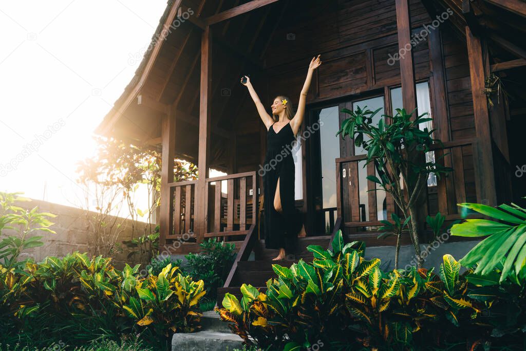 From below of exited female with outstretched arms standing near entrance in house while celebrating holiday on vacation in tropical country