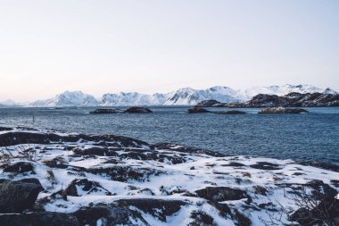 Norveç 'te kış zamanı bulutsuz mavi gökyüzüne karşı dalgalı denizin yakınında kayalık oluşumlarla karlı dağ sıraları