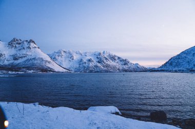 Kış zamanı alacakaranlıkta Norveç 'te bulutsuz gökyüzüne karşı dalgalanan denizin yakınında kayalık oluşumlarla karlı dağ sıraları