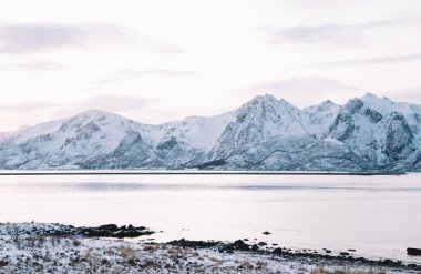 Bergsfjord köyünde karla kaplı kayalık dağlarla çevrili sakin deniz manzarası kışın bulutlu gün batımına karşı.