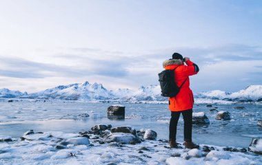 Sırt çantalı uzun bir gezgin, dürbünle bakışlarını kaçırarak vahşi dağlarla dolu soğuk, donmuş okyanus kıyısında duruyor.