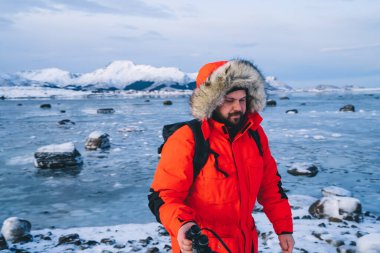 Dürbünlü ceketli sakallı adam Norveç 'te kayalar ve karla ıssız bir kıyıda tek başına yürürken.