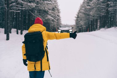 Norveç 'te kış mevsiminde ormanın yakınındaki karlı yolda dikilirken kollarını uzatıp araba yakalayan anonim bir insanın arka görüntüsü.