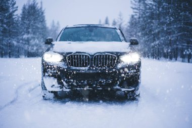 01 Şubat 2020: Pahalı siyah BMW otomobili, parlayan farları ile kış akşamları kar yağışı ve don altında her zaman yeşil ağaçların arasında sürüş yapıyor.