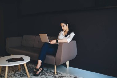 Tam vücut kendinden emin etnik kadın resmi olarak rahat koltukta oturuyor ve modern ofis alanında dizüstü bilgisayarla çalışıyor.