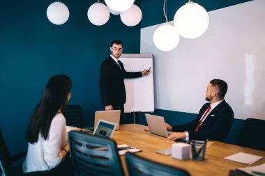 Resmi takım elbiseli ve gözlüklü konsantre genç erkek yönetici masada oturan ve dizüstü bilgisayarlar üzerinde çalışan meslektaşlarına çizimler gösteriyor.