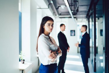 Resmi giysiler içindeki düşünceli bir bayanın ofis koridorunda gün ışığında konuşan meslektaşlarına karşı elleri çapraz duruşu.
