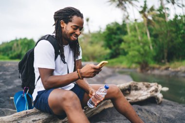 Yan görünüm içeriği Afrika kökenli Amerikalı erkek yürüyüşçü ağaç gövdesinde oturuyor ve tropikal ormanlarda dinlenirken cep telefonunu karıştırıyor.