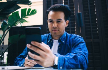 Kot giyen şık etnik erkek cep telefonu kullanıyor ve bitkinin yanında yapraklarla otururken video çekiyor.