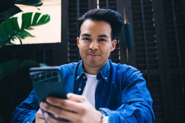 Modern konforlu iş yerinde kameraya bakıp cep telefonuyla mesajlaşan modaya uygun kot giymiş etnik bir erkek.