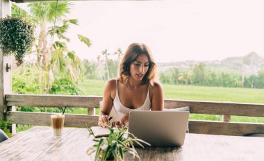 Kendine güveni tam, beyaz tişörtlü İspanyol bir kadın yaz günü açık bir kafede çalışırken dizüstü bilgisayarında video görüşmesi yapıyor.