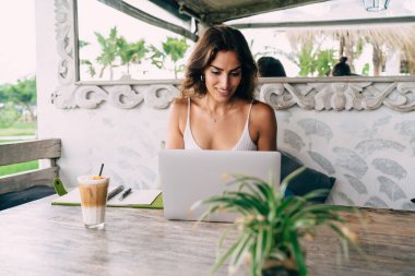 Modaya uygun kıyafetli genç bayan, elinde dizüstü bilgisayar ve bir bardak soğuk içecekle ahşap masada oturmuş tropikal kafe terasında uzaktan çalışıyor.