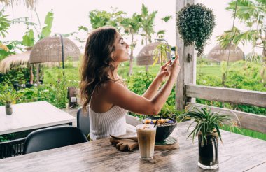 Yan görüş, beyaz bluz giyen genç bir bayanın tropikal bahçede egzotik yiyeceklerle masada otururken cep telefonuyla fotoğraf çekmesi.