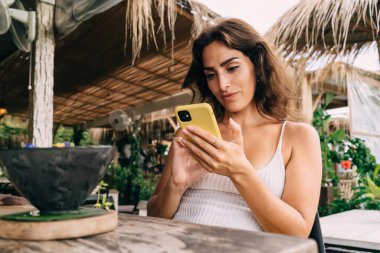 Güneşli bir günde, tropikal bir tatil köyünde egzotik yiyeceklerle masada otururken, üzerinde sarı kapaklı cep telefonuyla gezen iyimser genç bayan.