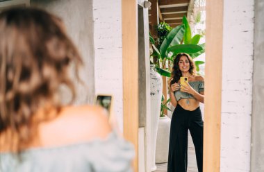 Modayı takip eden iyimser stil sahibi etnik kadın cep telefonuyla fotoğraf çekerken gülümseyip neşeyle aynaya bakıyor.