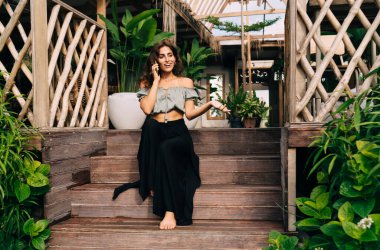 Şık giyinmiş, çıplak ayaklı bir kadın cep telefonuyla konuşuyor. Duygusal olarak el yayıyor. Güneşli bir günde bitkilerin arasında dinlenen, ağaçtan yapılmış bir terasın merdivenlerinde oturuyor.