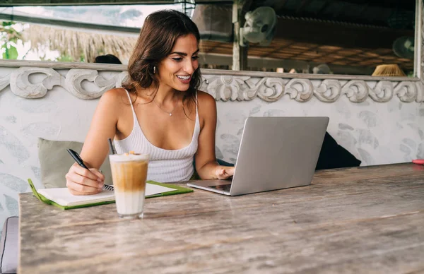 夏の服で積極的なヒスパニック系の女性は ノートパソコンを使用して アイスコーヒーのガラスと居心地の良い屋外カフェでリモートで作業しながらノートを取ります — ストック写真