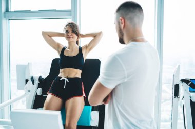 Kollarını kavuşturmuş, anonim sakallı antrenör. Gülümseyen sporcu kadının yanında. Ellerini kafasının arkasına koyup spor salonundaki modern ekipman üzerinde çalışıyor.